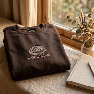 May include: A dark brown crewneck sweatshirt featuring an embroidered design of a loaf of bread centered on the chest. Below the bread illustration, the text "wake and bake" is embroidered in a light beige thread in a simple, clean font.
