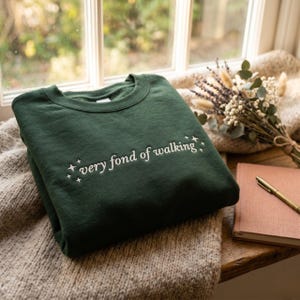 Puede incluir: Una sudadera de cuello redondo en color verde bosque oscuro con el texto bordado en blanco "very fond of walking" centrado en el pecho, flanqueado por pequeños iconos de estrellas de cuatro puntas a ambos lados de las letras.