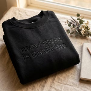 Op de afbeelding: Zwart sweatshirt met ronde hals met de geborduurde tekst "movement is medicine". Het sweatshirt is netjes op een neutraal gekleurd oppervlak gevouwen. Een notitieboek, potlood en takje gedroogde bloemen staan op de achtergrond.