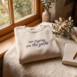 May include: A beige crewneck sweatshirt featuring the dark blue embroidered text "no crying on the yacht" centered on the chest. The garment has long sleeves, a ribbed neckline, and a smooth fabric finish, presented in a folded, flat orientation.