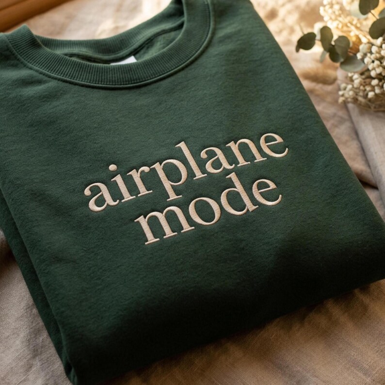 May include: A dark green sweatshirt with the words "airplane mode" embroidered in a light beige color. The sweatshirt has a crew neck and a relaxed fit, perfect for casual wear.