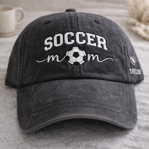 May include: A charcoal gray baseball cap with the words "SOCCER MOM" embroidered in white, along with a soccer ball graphic. The name "TAYLOR" is embroidered on the side. The cap has a curved brim and a top button.