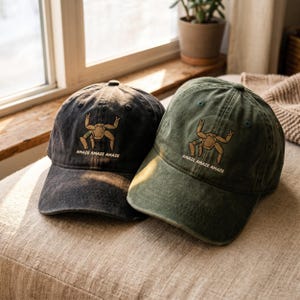 May include: Two baseball caps, one in charcoal gray and one in olive green, both featuring an embroidered graphic of a tan, multi-legged creature with raised arms and the text "AMAZE AMAZE AMAZE" stitched in white thread below the design.