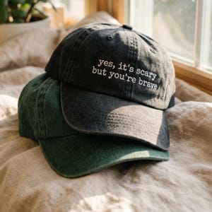 Può includere: Un cappellino da baseball grigio antracite con il testo ricamato in bianco yes, it's scary but you're brave, sovrapposto a un cappellino da baseball verde scuro. Entrambi i cappelli presentano una texture in tessuto slavato e visiera curva.