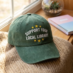 Peut inclure: Casquette de baseball verte avec le texte "SUPPORT YOUR LOCAL LIBRARY" en blanc et des étoiles jaunes. Le chapeau est posé sur un pull beige, avec une fenêtre et un carnet en arrière-plan.