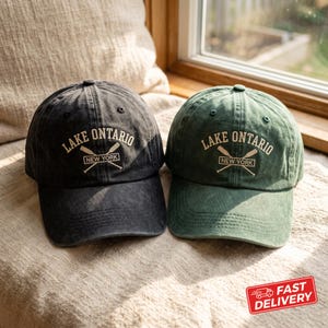 Könnte beinhalten: Zwei Baseballkappen, eine in Anthrazit und eine in Waldgrün, beide mit gesticktem Text LAKE ONTARIO, einer Grafik mit gekreuzten Rudern und dem Wort NEW YORK in einem rechteckigen Rahmen auf den vorderen Paneelen versehen.