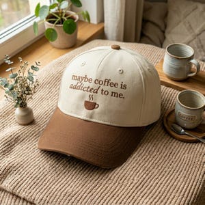 May include: A beige and brown baseball cap with the embroidered text "maybe coffee is addicted to me." A brown coffee cup graphic is below the text. The cap has a curved brim and a button on top. The cap is on a tan blanket.