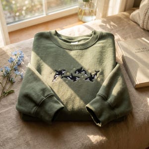 Könnte beinhalten: Ein olivgrünes Sweatshirt mit gestickten Orca-Walen und Sternen. Das Kleidungsstück ist ordentlich auf einer neutralen Oberfläche gefaltet, mit einem Buch und Blumen im Hintergrund. Das Design zeigt schwarz-weiße Orcas.