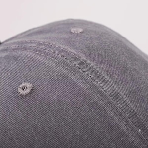May include: Close-up of a gray baseball cap. The cap is made of a textured fabric and features a curved brim. The cap has a stitched seam and a small, embroidered ventilation hole.