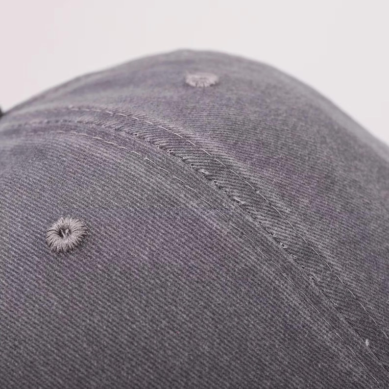 May include: Close-up of a gray baseball cap. The cap is made of a textured fabric and features a curved brim. The cap has a stitched seam and a small, embroidered ventilation hole.