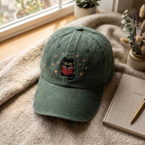 May include: A green baseball cap with an embroidered design of a black cat wearing glasses and reading a red book, surrounded by gold stars. The cap is sitting on a beige blanket near a window.