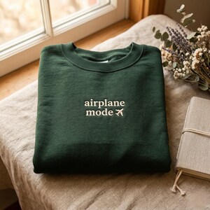 May include: A dark green sweatshirt with the words "airplane mode" embroidered in white, along with a small airplane graphic. The sweatshirt is folded and resting on a neutral-colored surface, with a window and dried flowers in the background.
