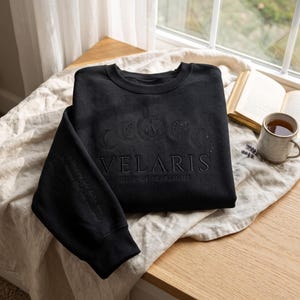 以下が含まれることがあります： 月の満ち欠けの刺繍と「VELARIS CITY OF STARLIGHT」の文字が施された黒いクルーネックのスウェットシャツ。袖には「To the stars who listen and the dreams that are answered.」という刺繍が施されています。