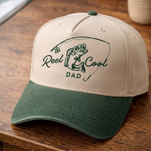 May include: A beige baseball cap with a dark green brim and a dark green graphic print on the front. The graphic features a fishing rod, a fish, and the text Reel Cool DAD. The cap has a structured crown and a small green button on top.