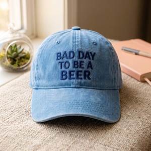 May include: A light blue denim baseball cap with a six-panel construction and a curved brim. The front of the cap features dark blue embroidered text that reads "BAD DAY TO BE A BEER" in a bold, sans-serif font. The fabric has a washed texture.