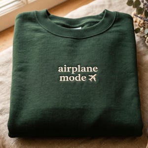 May include: A dark green sweatshirt with the words "airplane mode" embroidered in white, along with a small airplane icon. The sweatshirt is folded neatly on a neutral-colored surface.