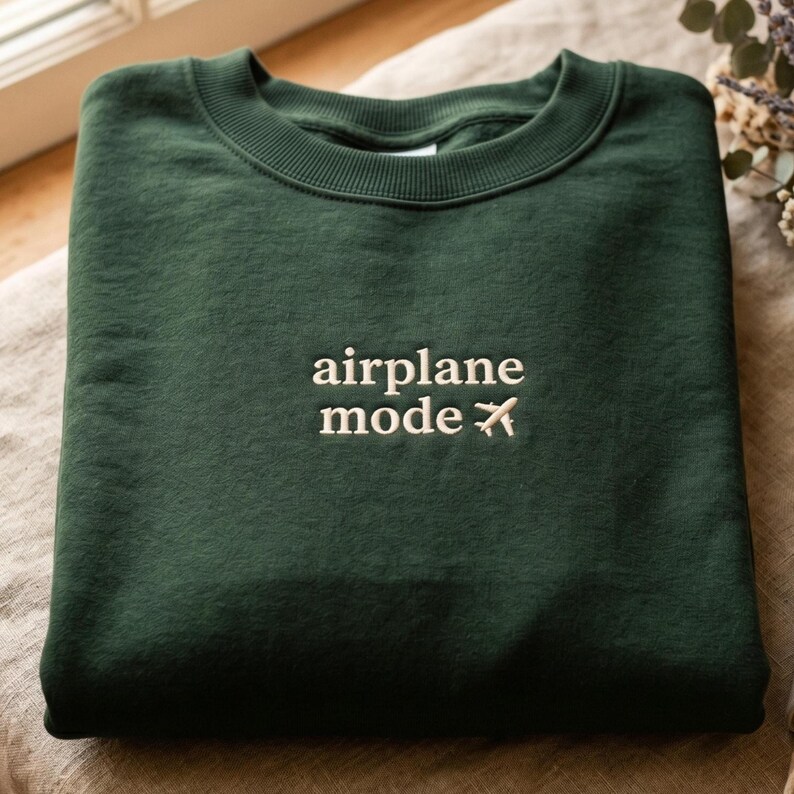 May include: A dark green sweatshirt with the words "airplane mode" embroidered in white, along with a small airplane icon. The sweatshirt is folded neatly on a neutral-colored surface.