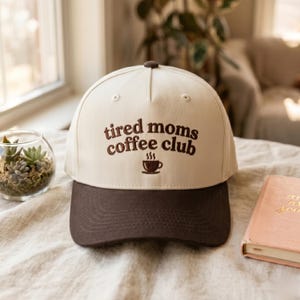 May include: A cream-colored baseball cap with a dark brown brim. The front features the embroidered text "tired moms coffee club" in dark brown, positioned above a small, matching dark brown embroidered icon of a steaming coffee cup.