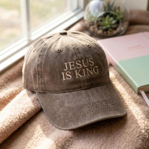 Könnte beinhalten: Braune Baseballkappe mit dem gestickten Text "JESUS IS KING" in hellem Beige. Die Kappe liegt auf einer weichen, strukturierten Oberfläche in der Nähe eines Fensters. Im Hintergrund ist ein rosa-grünes Buch zu sehen.
