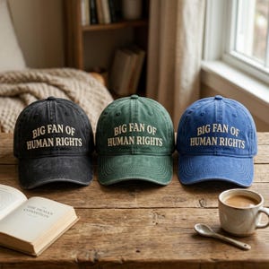 May include: Three baseball caps in black, green, and blue, each with the text "BIG FAN OF HUMAN RIGHTS" in white. The hats are displayed on a wooden surface with a book and a coffee cup.
