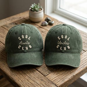 May include: Two olive green baseball caps with the text "COOL AUNTS CLUB" and "COOL UNCLES CLUB" embroidered in white. The caps are displayed on a wooden surface, with a small potted plant and decorative stones in the background.