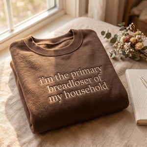 May include: A brown sweatshirt with the embroidered text "I'm the primary breadloser of my household." The crew neck sweatshirt is folded and resting on a light-colored surface. A notebook and pen are visible in the background.
