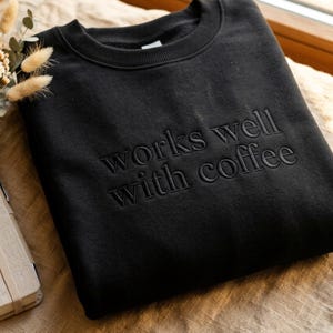 May include: Black crew neck sweatshirt with the phrase "works well with coffee" embroidered in a tone-on-tone color. The sweatshirt is made of a soft material and is displayed on a neutral surface.