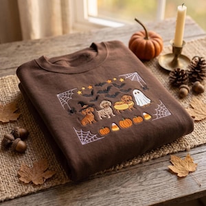 May include: A brown crewneck sweatshirt featuring an embroidered Halloween design. The embroidery includes three dachshunds dressed as a witch, a mummy, and a hot dog, alongside a ghost, bats, stars, spiderwebs, pumpkins, and candy corn pieces.
