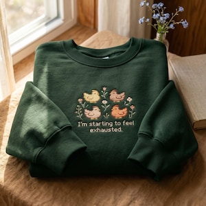 Op de afbeelding: Een donker bosgroene sweater met ronde hals voorzien van een borduursel van vier kleine, kleurrijke vogels omringd door kleine bloemetjes. Onder de vogels is de tekst "I'm starting to feel exhausted." geborduurd in een eenvoudig, strak lettertype.