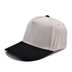 May include: A baseball cap featuring a light beige crown and a contrasting solid black visor. The cap is constructed with a structured five-panel design, reinforced stitching along the brim, and a small black button detail at the top center.