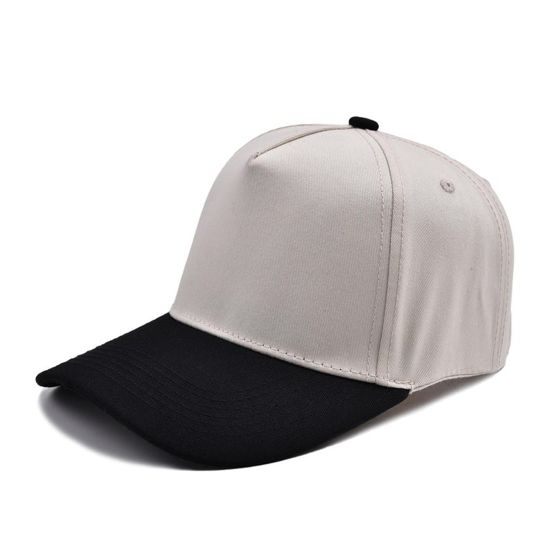 May include: A baseball cap featuring a light beige crown and a contrasting solid black visor. The cap is constructed with a structured five-panel design, reinforced stitching along the brim, and a small black button detail at the top center.
