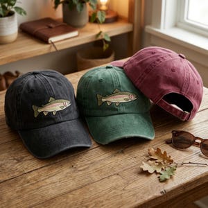 Op de afbeelding: Drie baseballpetten in zwart, groen en bordeauxrood, elk met een vispatch. De petten liggen op een houten oppervlak met zonnebril en bladeren. De petten hebben een distressed look.