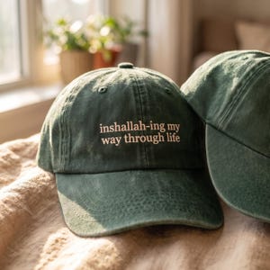 May include: A forest green baseball cap with a curved brim and an adjustable strap. The front of the cap features embroidered white text that reads, "inshallah-ing my way through life," centered above the brim in a simple, clean font style.