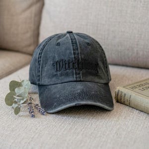 May include: A dark gray, distressed baseball cap with the word "Witching" embroidered on the front. The hat is sitting on a neutral-toned surface, with a sprig of eucalyptus and a vintage book nearby.