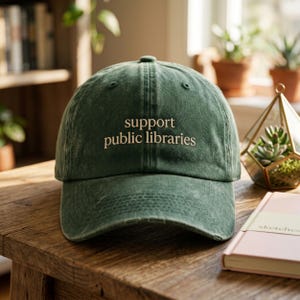 May include: A dark green baseball cap with the embroidered text "support public libraries" in a light beige color. The cap is sitting on a wooden surface. The hat has a curved brim and a classic six-panel design.