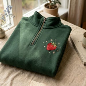 May include: A dark green sweatshirt with a quarter-zip neckline and a red embroidered strawberry design. The strawberry is surrounded by small embroidered stars. The sweatshirt is folded neatly on a light-colored surface.
