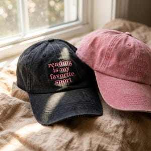 May include: Two baseball caps, one charcoal black and one dusty rose pink, both featuring the embroidered text reading is my favorite sport in pink lettering. Each cap has a six-panel construction with a curved brim and a distressed fabric finish.
