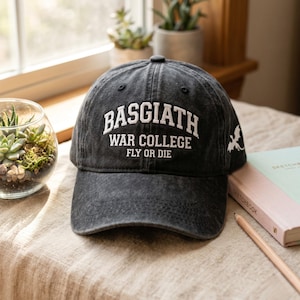 以下が含まれることがあります： 「BASGIATH WAR COLLEGE FLY OR DIE」の白文字が入った黒い野球帽。ベージュの表面に置かれ、側面に小さなドラゴンのグラフィックがあります。鉛筆とスケッチブックも表示されています。