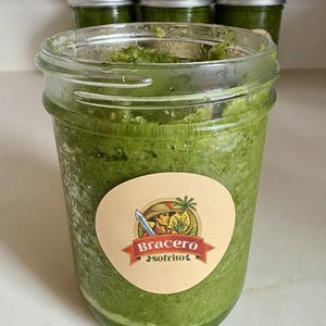 May include: A clear glass jar filled with vibrant green sofrito, with a label that reads "Bracero sofrito". Several other jars of sofrito are visible in the background, all with silver lids.