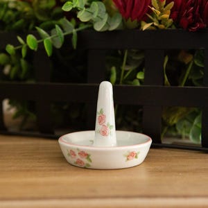 May include: A white ceramic ring holder with a floral rose design. The ring holder has a small dish and a central post for holding rings. The dish and post are decorated with pink roses and green leaves.