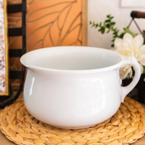 May include: A white ceramic chamber pot with a smooth, glossy finish, a wide circular opening, and a single curved handle on the side. The vessel features a rounded, bulbous body shape and a slightly flared rim, presenting a clean and simple design.