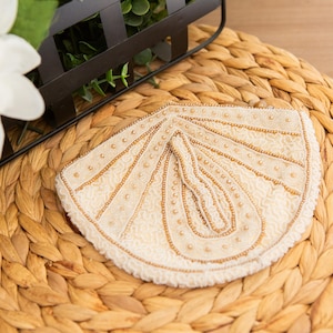 May include: A semi-circular cream-colored fabric applique featuring intricate gold beadwork and embroidery in a fan-like pattern. The design includes radiating lines of small gold beads and a central teardrop shape, all set against a textured lace base.