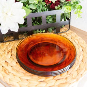 May include: A stack of four round amber-colored glass plates with a smooth, translucent finish. The plates feature a simple, circular design with a slightly raised rim, showcasing a deep, warm orange hue that catches the light across the surface.