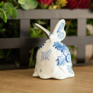 May include: A white ceramic bell with blue floral and geometric patterns, topped with a butterfly-shaped handle. The bell is set on a wooden surface, with a dark metal basket and greenery in the background.