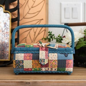 May include: A small, blue patchwork sewing box with a matching handle. The box is decorated with a variety of colorful fabric squares and a patterned bow closure. The box is sitting on a wooden surface.