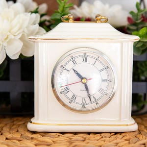May include: A cream-colored ceramic mantel clock with a rectangular body, a gold-trimmed base and top, and a gold handle. The clock face features black Roman numerals, a silver bezel, black hour and minute hands, and a red second hand.