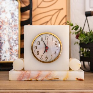 May include: A square, white marble clock with a gold-colored bezel and black numerals. The clock sits on a rectangular marble base with pink and yellow veining. Two cylindrical marble accents flank the clock.