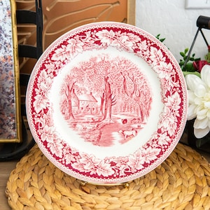 May include: A round ceramic plate featuring a red and white transferware pattern. The center depicts a rural landscape with a house, trees, and animals, while the wide rim is decorated with a detailed border of oak leaves and acorns.
