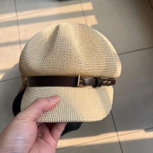 May include: A beige woven hat with a dark brown leather band and a gold-colored buckle. The hat has a rounded crown and a small brim. The hat is held by a hand.