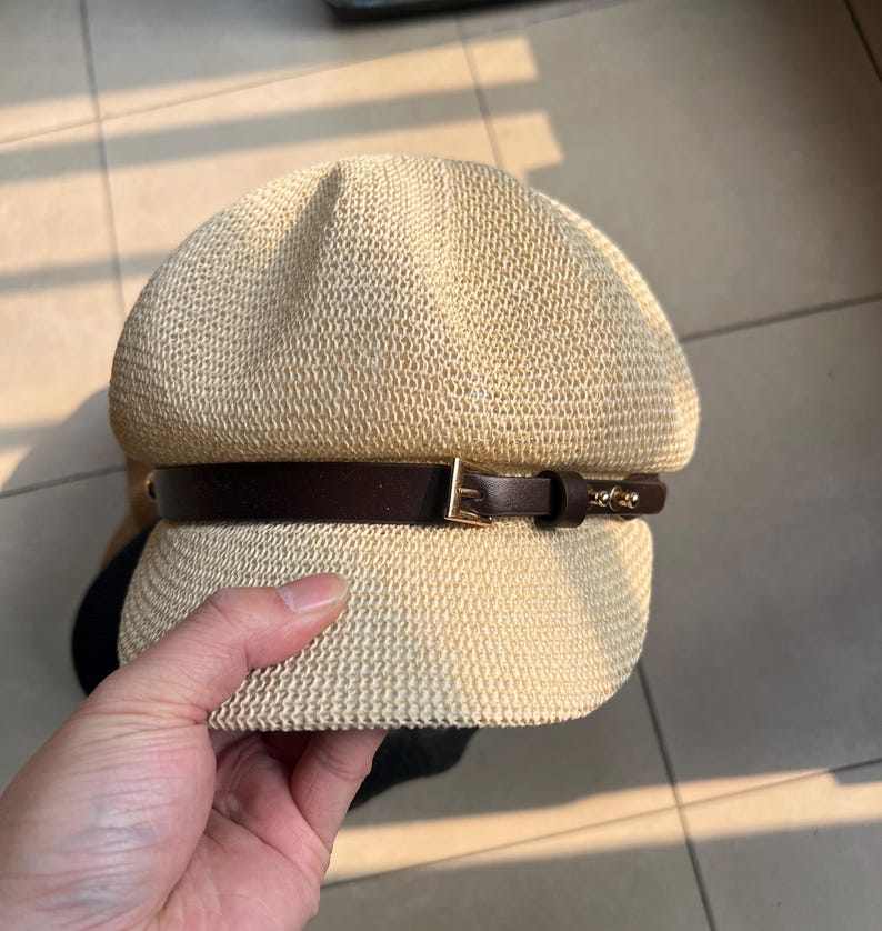 May include: A beige woven hat with a dark brown leather band and a gold-colored buckle. The hat has a rounded crown and a small brim. The hat is held by a hand.